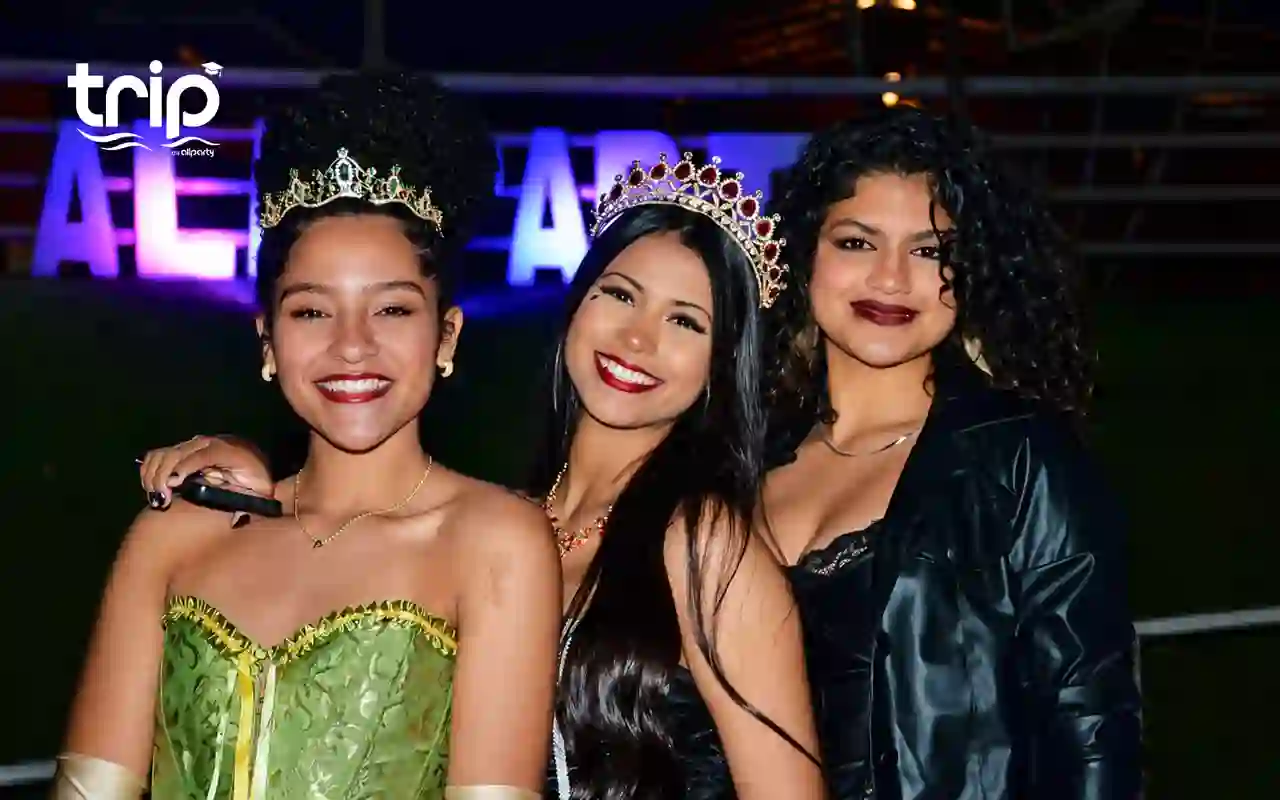 Três formandas com tiaras de festa, com o letreiro da All Party iluminado ao fundo